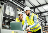 Two people in hard hats and yellow vests looking at a computer in a warehouse.